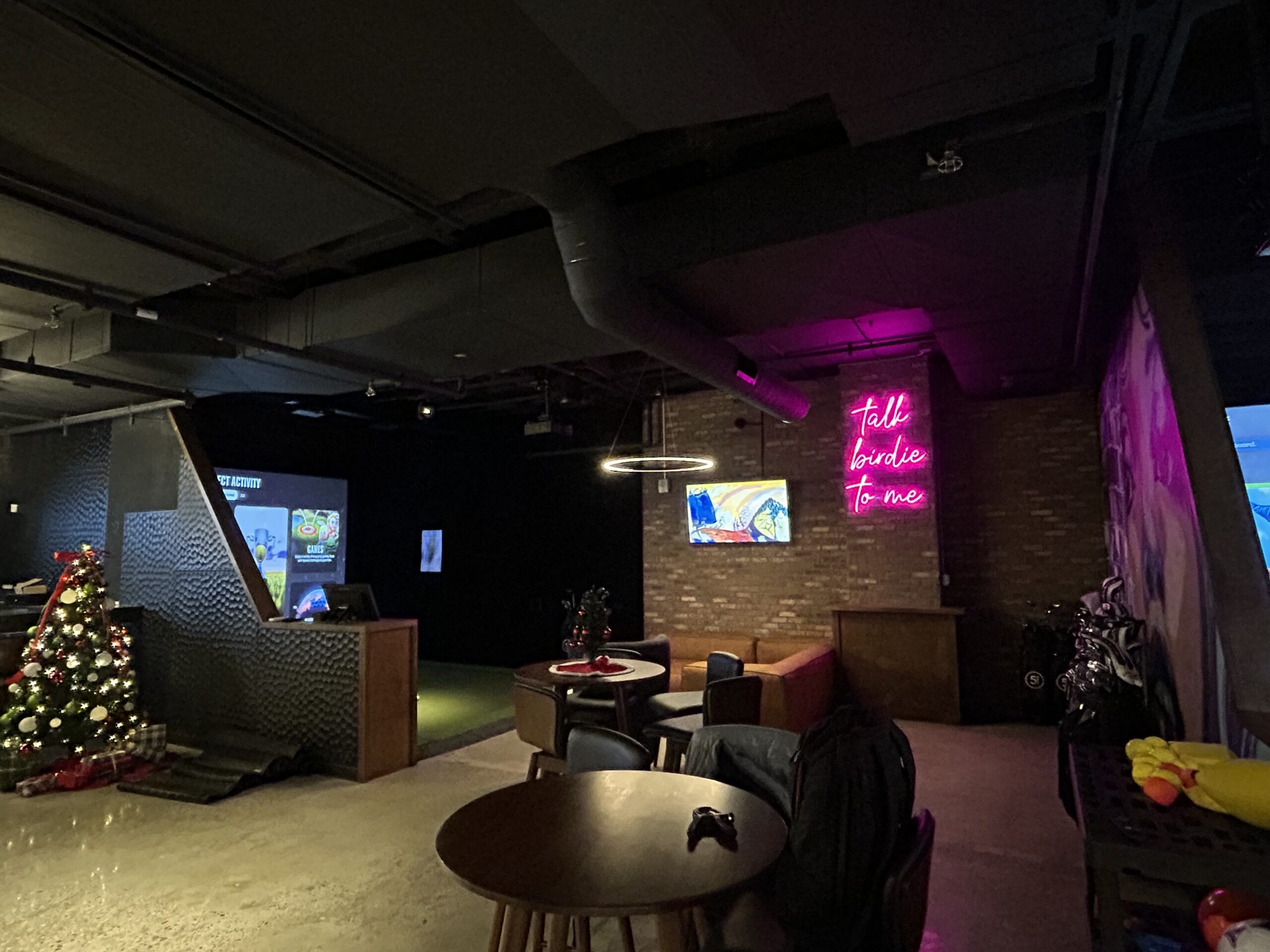 Dimly lit lounge with a round table, seating area, neon sign reading Talk birdie to me, Christmas tree, and golf simulator screens in the background