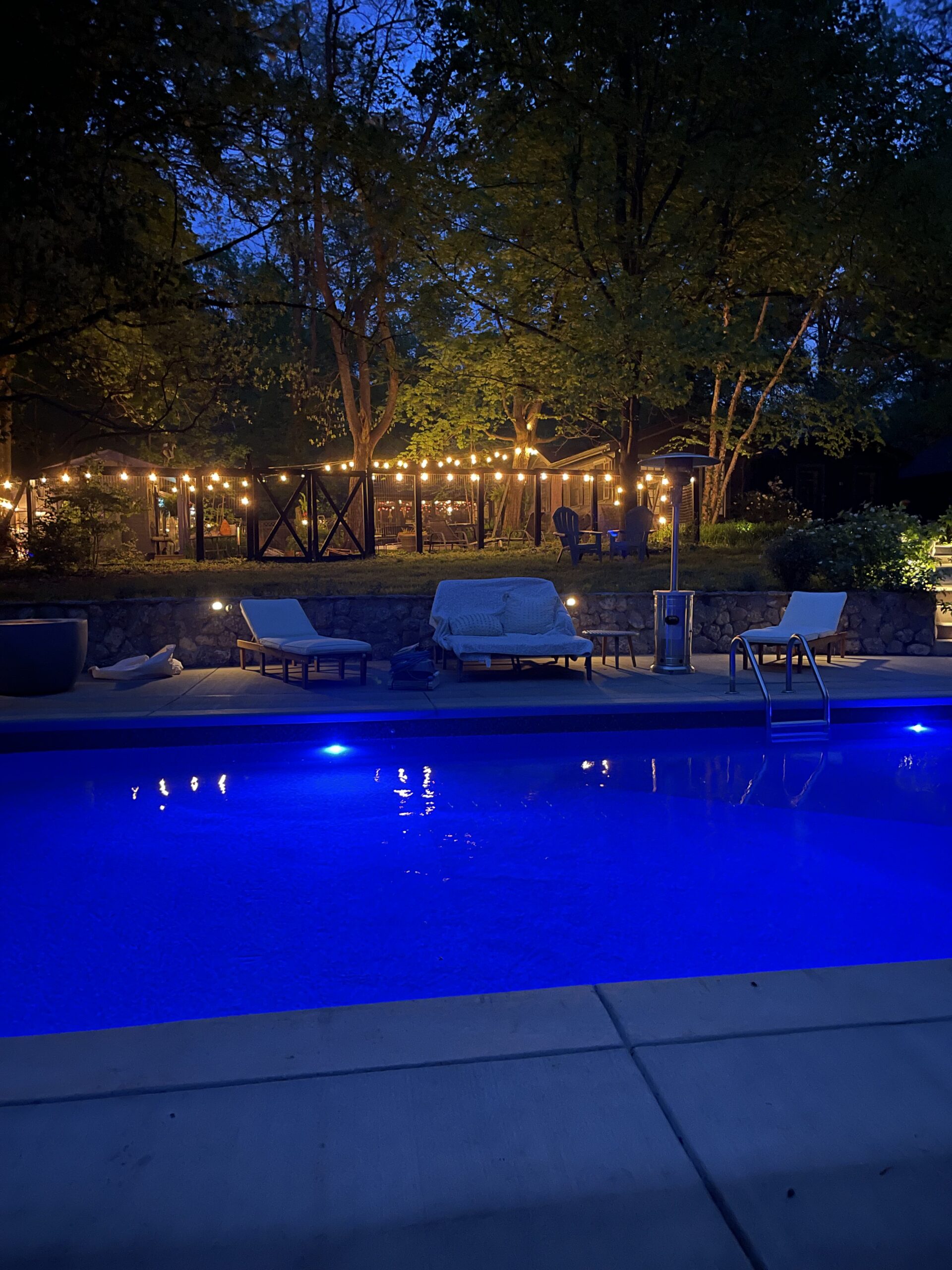 A swimming pool lit with blue lights at night, with lounge chairs poolside and string lights illuminating a patio and trees in the background.