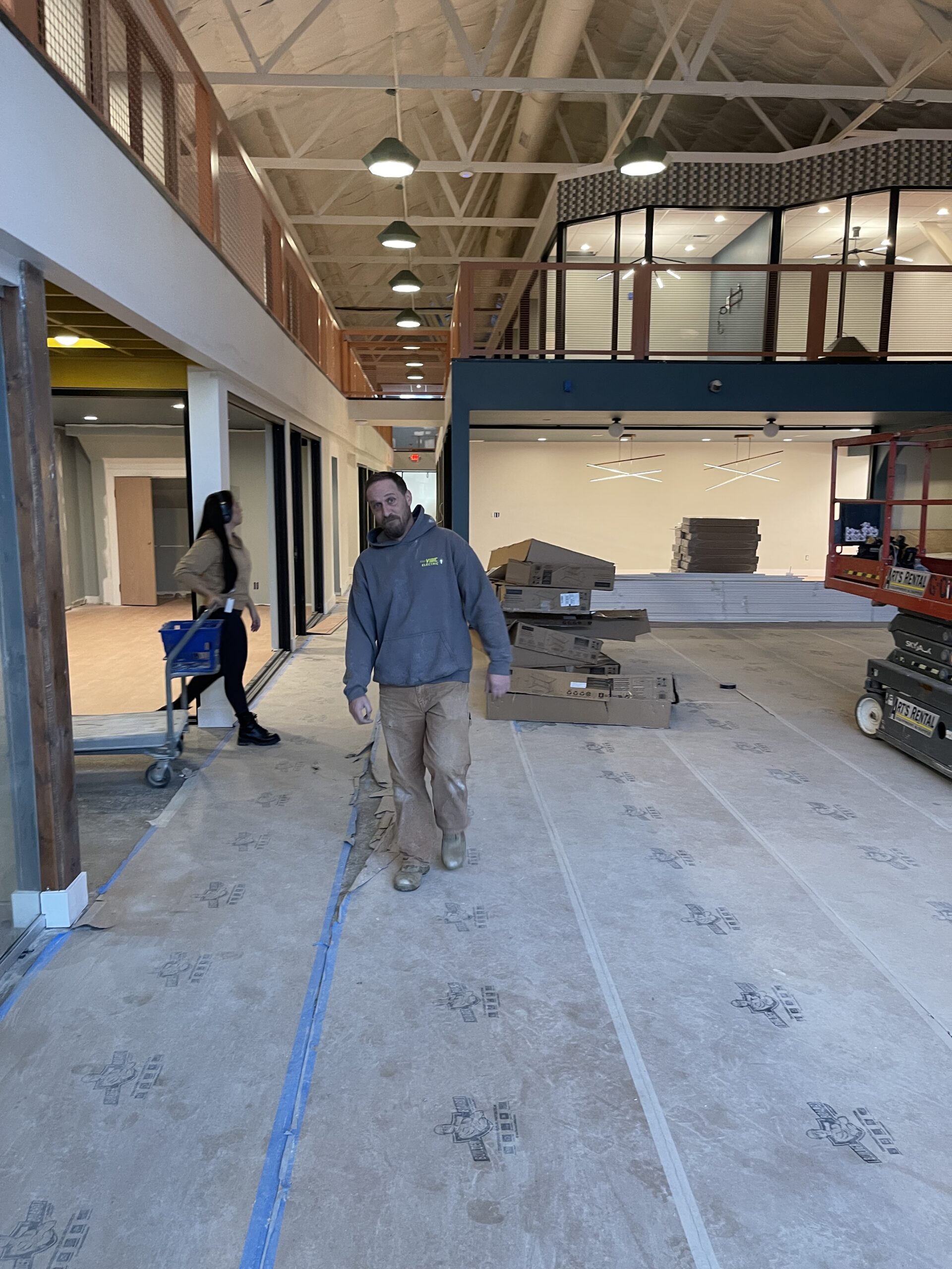 A man in work clothes walks through a construction site with labeled boxes and equipment; a woman in the background pushes a cart down a hallway.