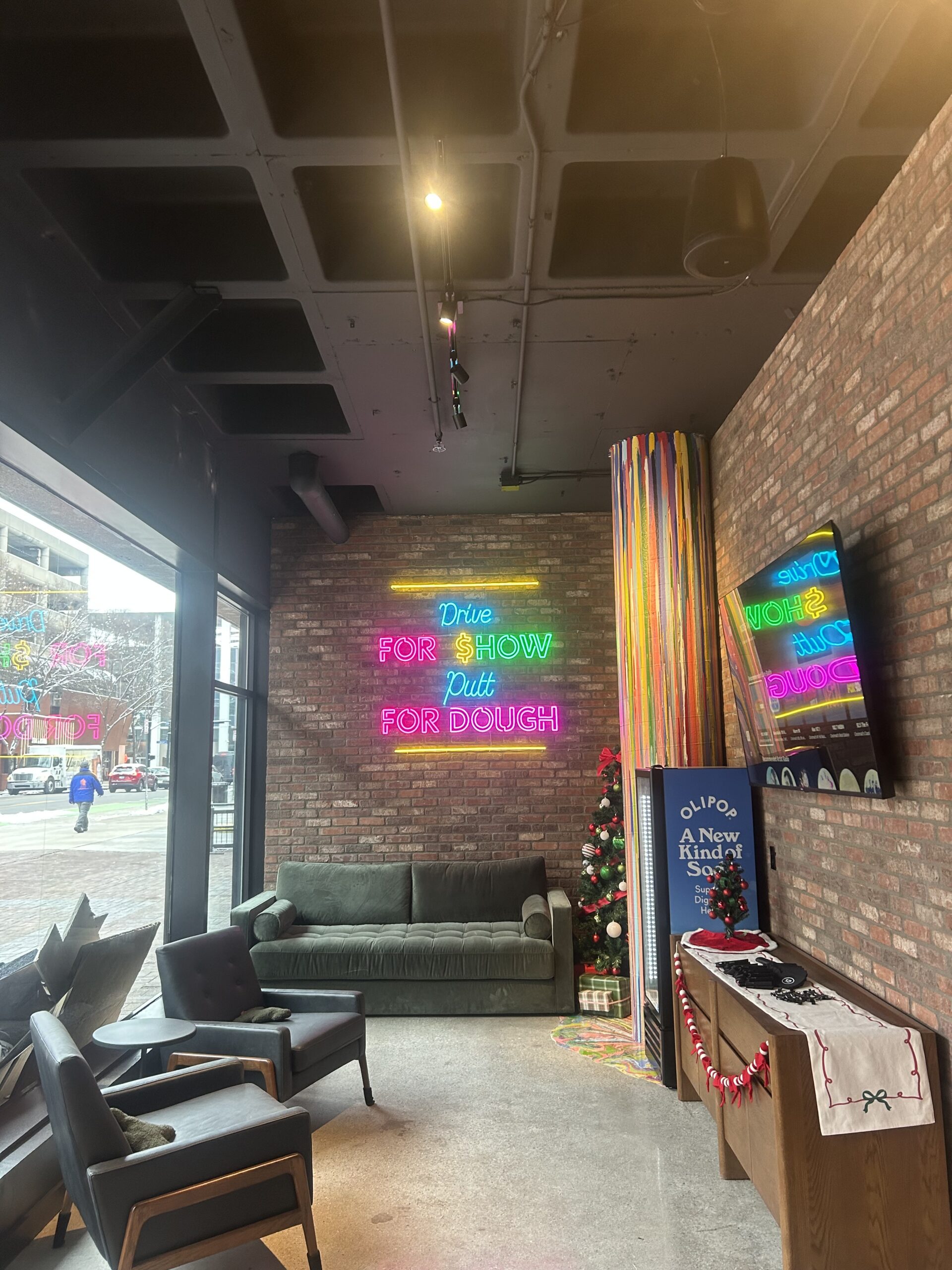 A modern lounge with brick walls, neon signs reading Drive FOR SHOW, Putt FOR DOUGH, a green sofa, a TV, and a decorated Christmas tree near the window