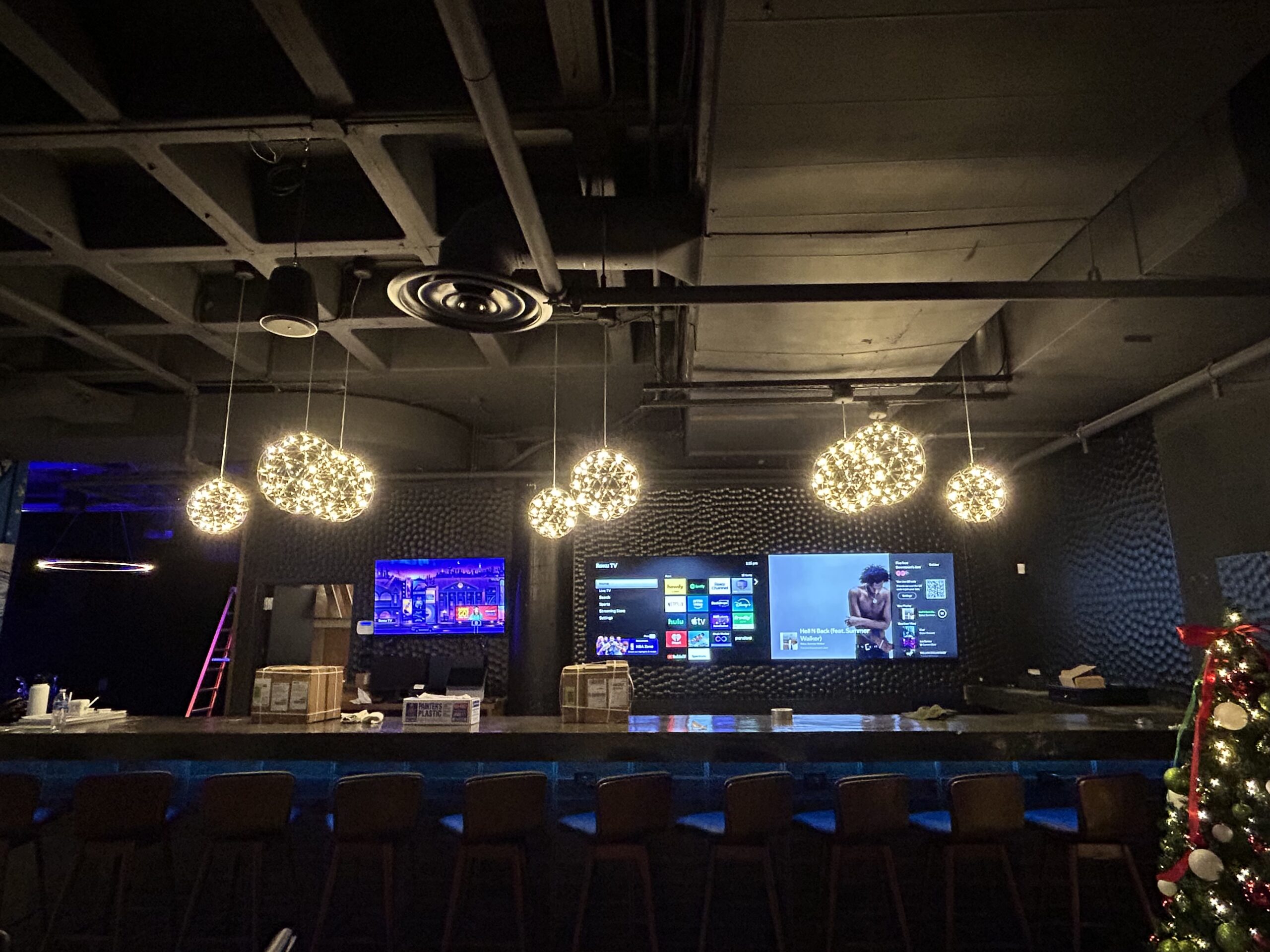 A modern bar with spherical pendant lights, a long counter with stools, large TV screens on the back wall, and a decorated Christmas tree on the right.