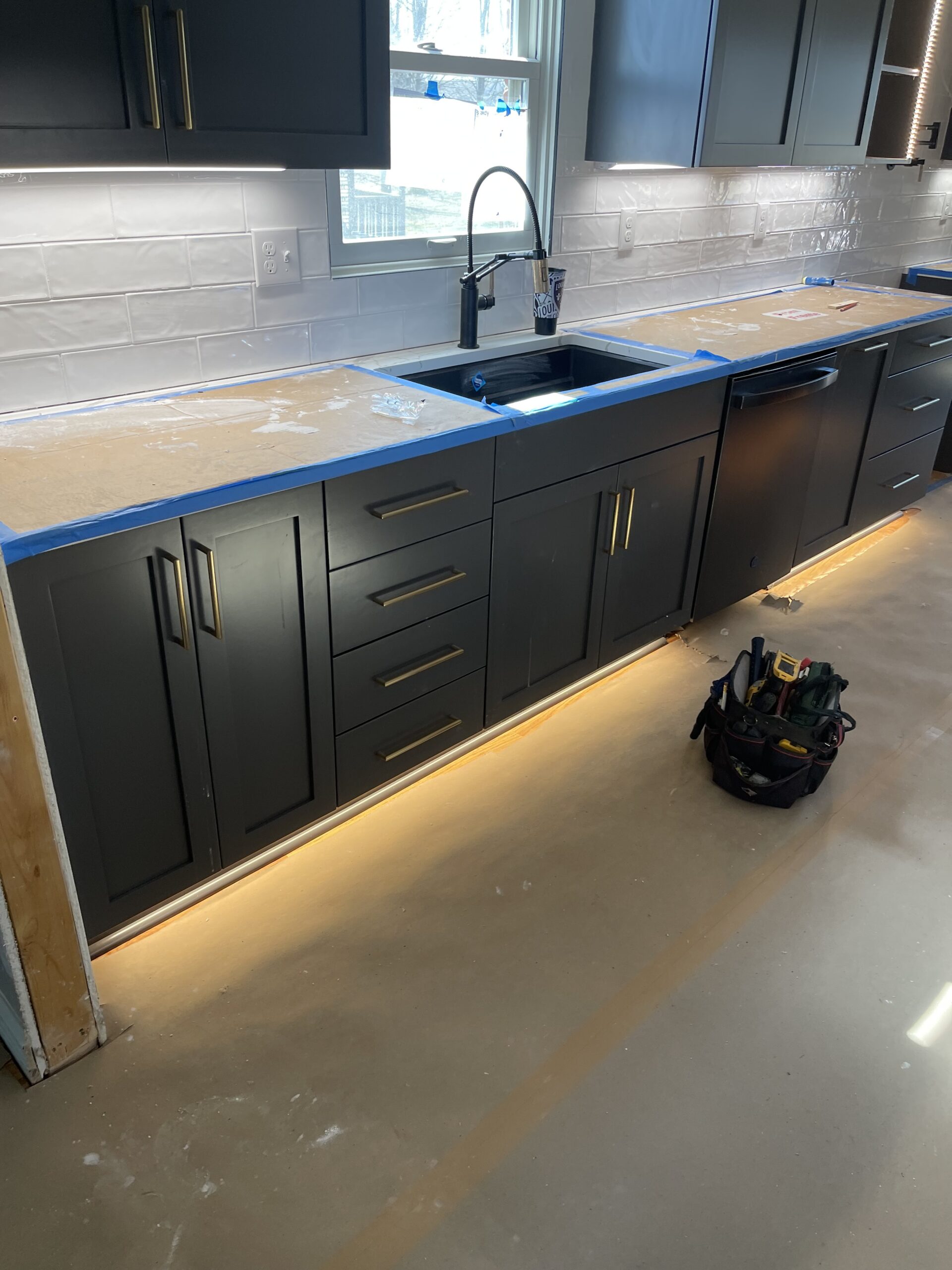 A kitchen under renovation with dark cabinets, missing countertops, tools on the floor, and under-cabinet lighting turned on.