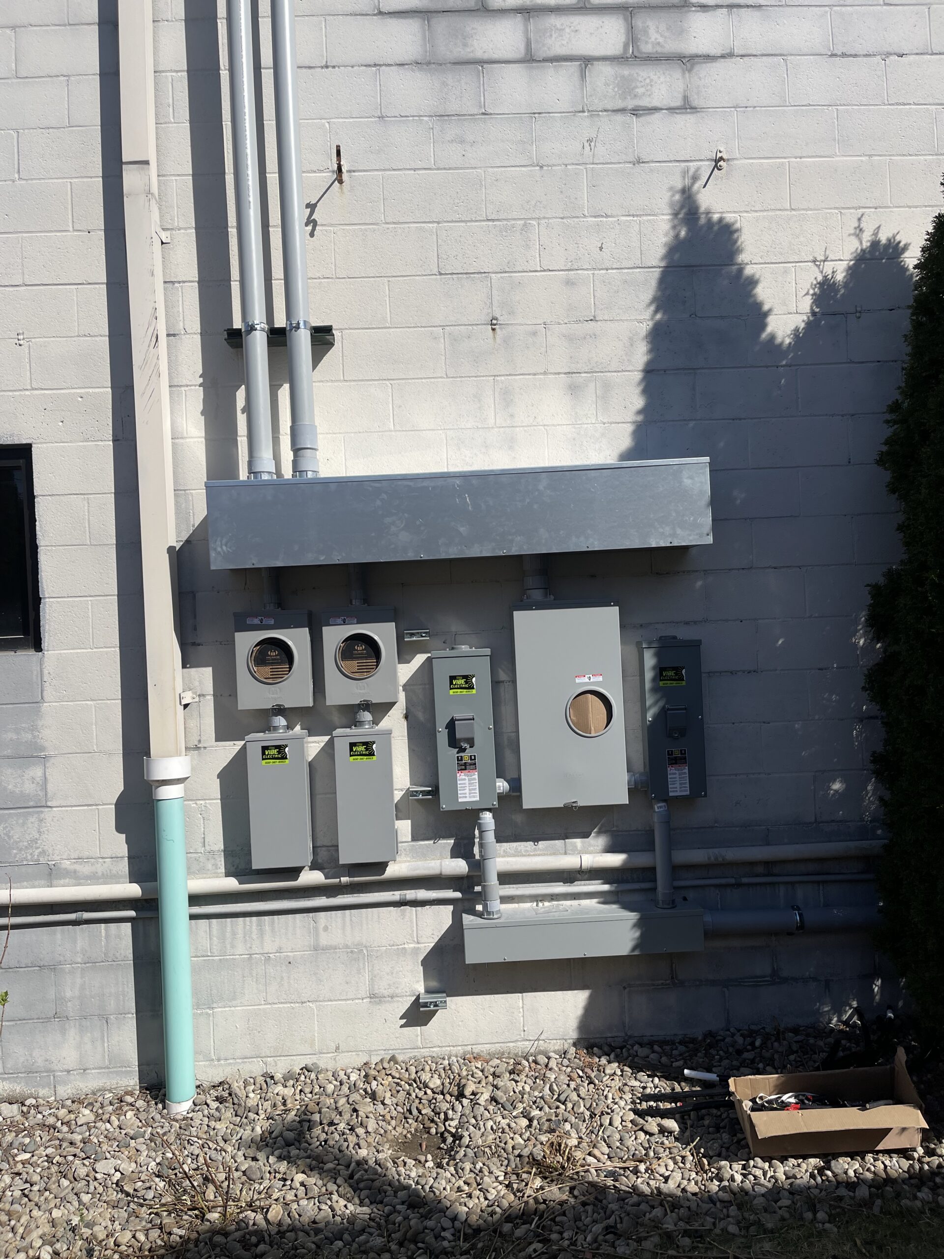 Electrical meters and panels mounted on an exterior cinderblock wall, with conduit pipes running vertically and horizontally; some sunlight and tree shadow visible.
